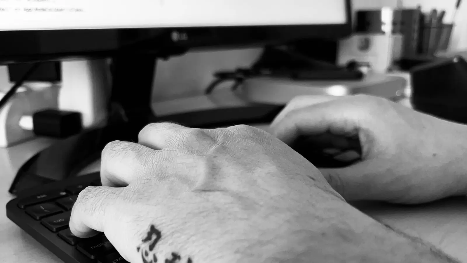 Una mano tatuata sulla tastiera davanti a un monitor, in bianco e nero.